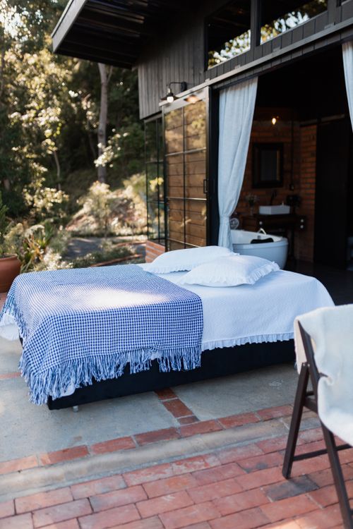 Cama matrimonial al aire libre bajo techo, con colcha azul a cuadros y funda blanca, junto a cortinas blancas y ambiente de dormitorio pequeño. Aprovechar espacio habitacion matrimonio pequeña.