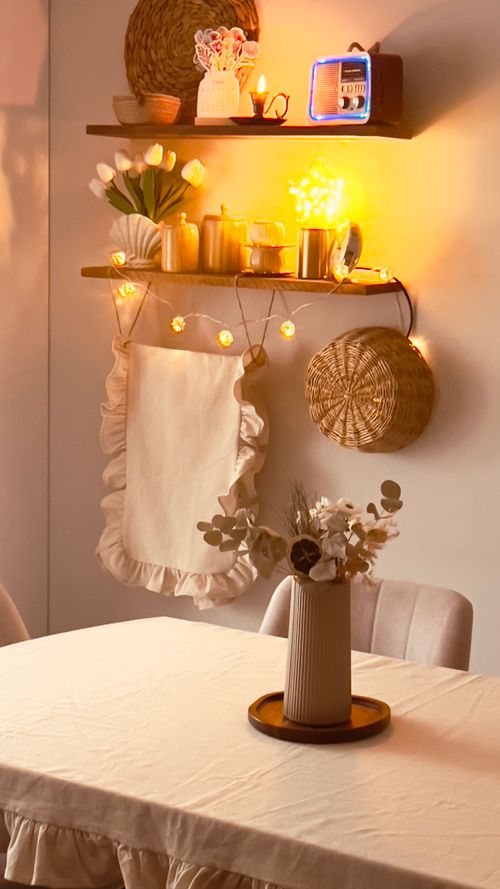 Mesa de comedor con mantel claro, jarrón floral en centro y luminarias cálidas; estantería decorativa con objetos, toallas colgantes y cesta de palma en pared. Integrar: últimas tendencias en decoración de dormitorios de matrimonio.