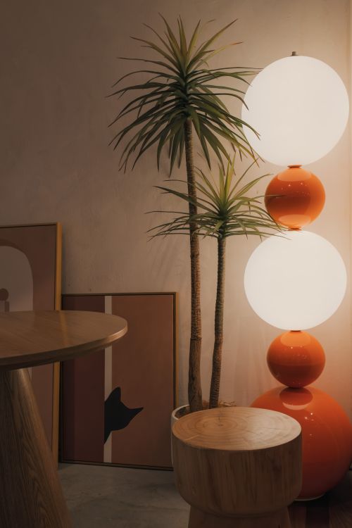 Salón minimalista con plantas altas y lámparas de bolos anaranjados; muebles de madera y cuadros apoyados en el suelo.