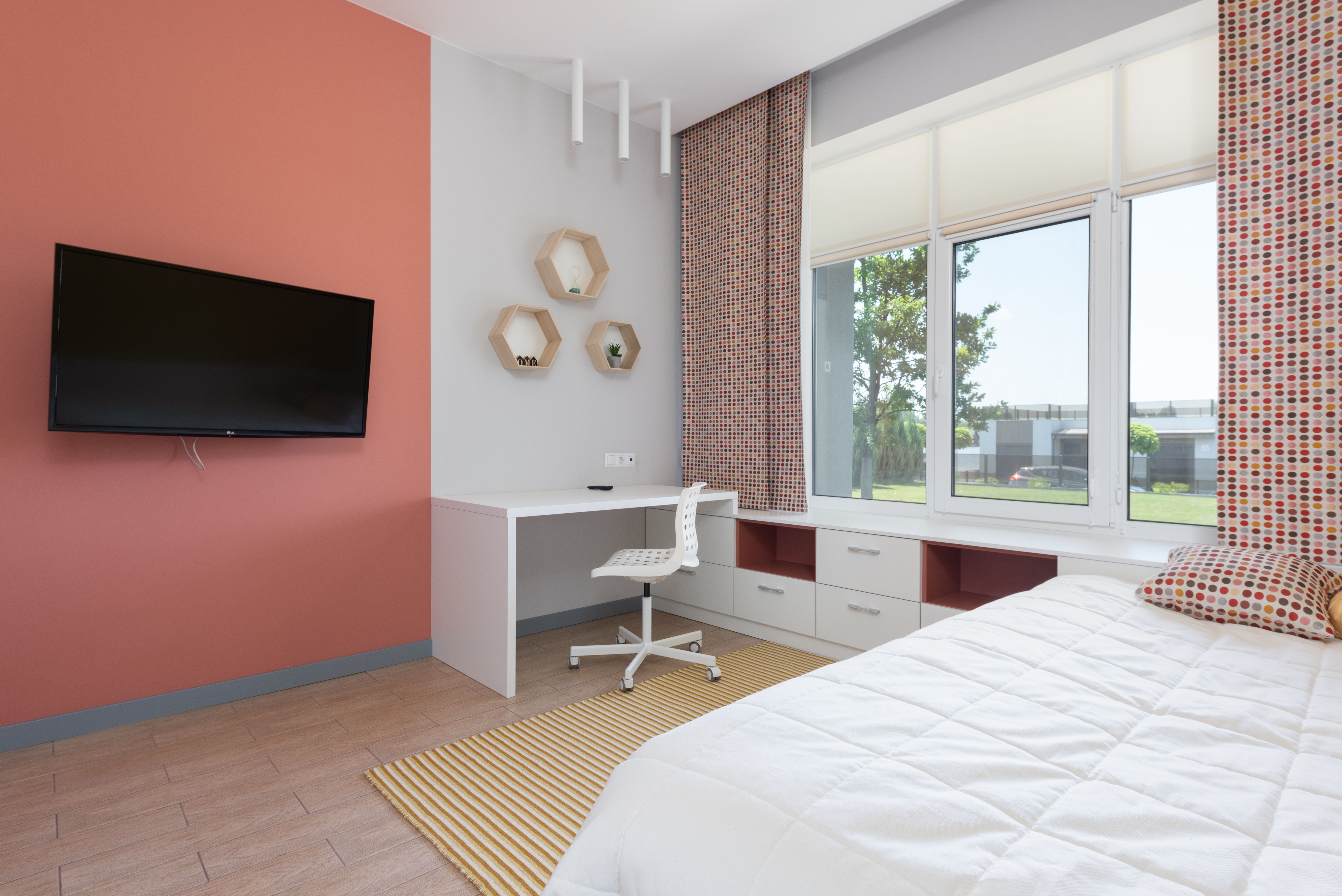 Habitación juvenil con cama, escritorio y televisión en una pared naranja, y gran ventanal que deja entrar la luz.