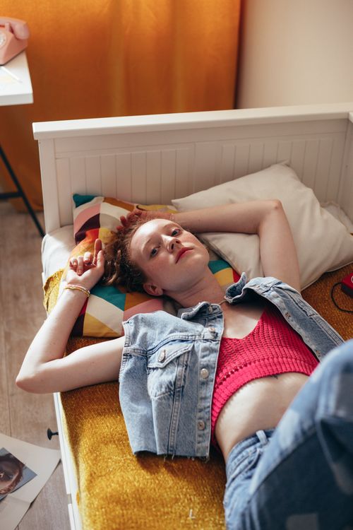 Una joven tumbada en la cama con chaqueta vaquera y top rosa, rodeada de cojines coloridos.