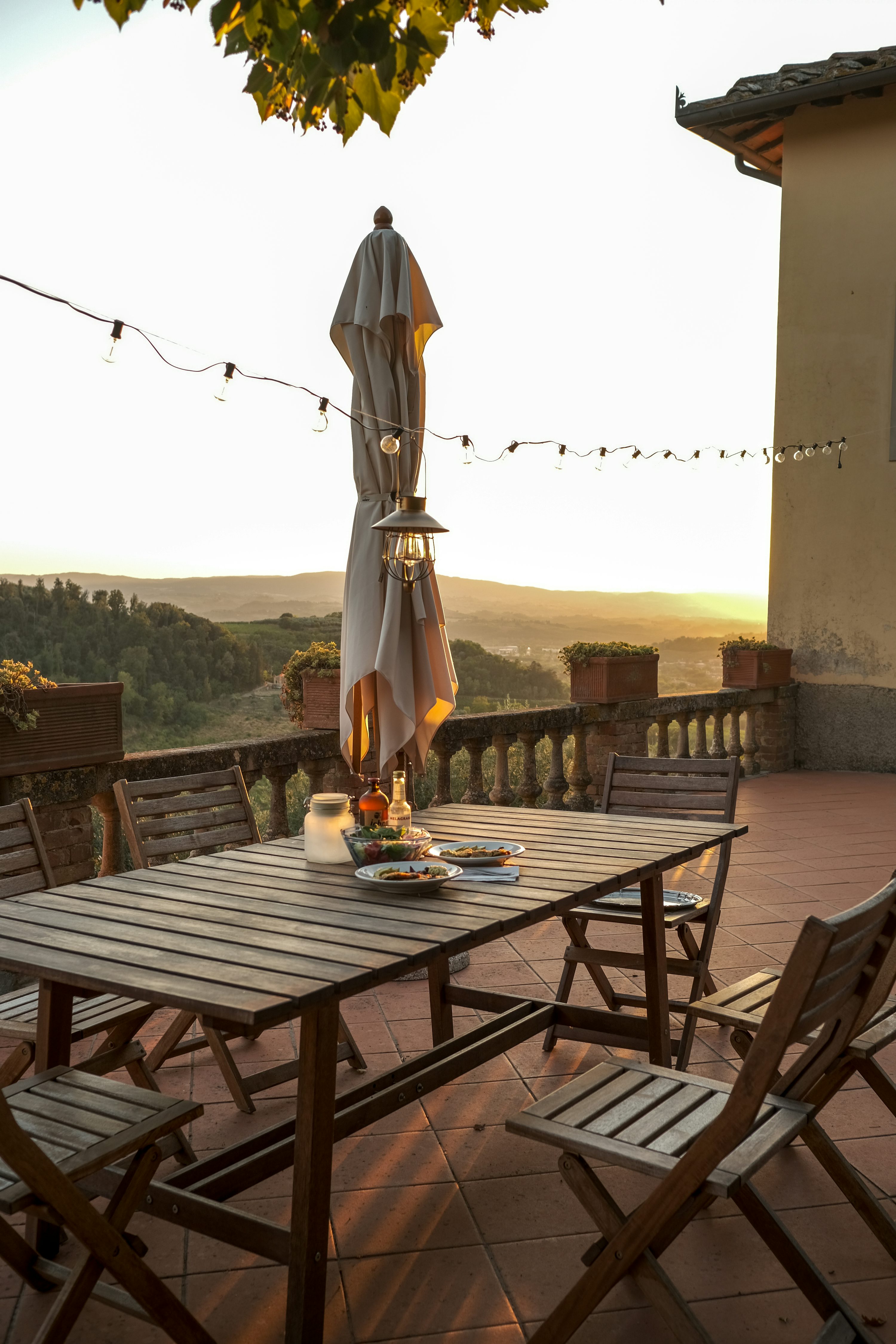 Mesa de exterior con sillas de madera y paraguas plegado en terraza al atardecer.