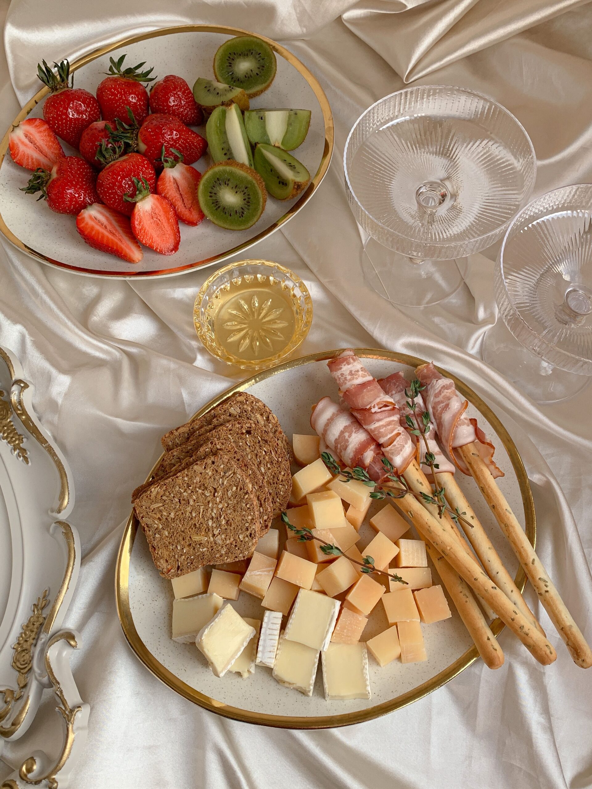 Plato de aperitivos con quesos y pan, acompañado de tiras de bacon enrolladas y palitos de pan; ensalada de fresas y kiwis, copas y vaso decorativos sobre una mantilla satinada. Incluye el contexto de “tamaño ideal de una mesa de comedor” en la descripción.