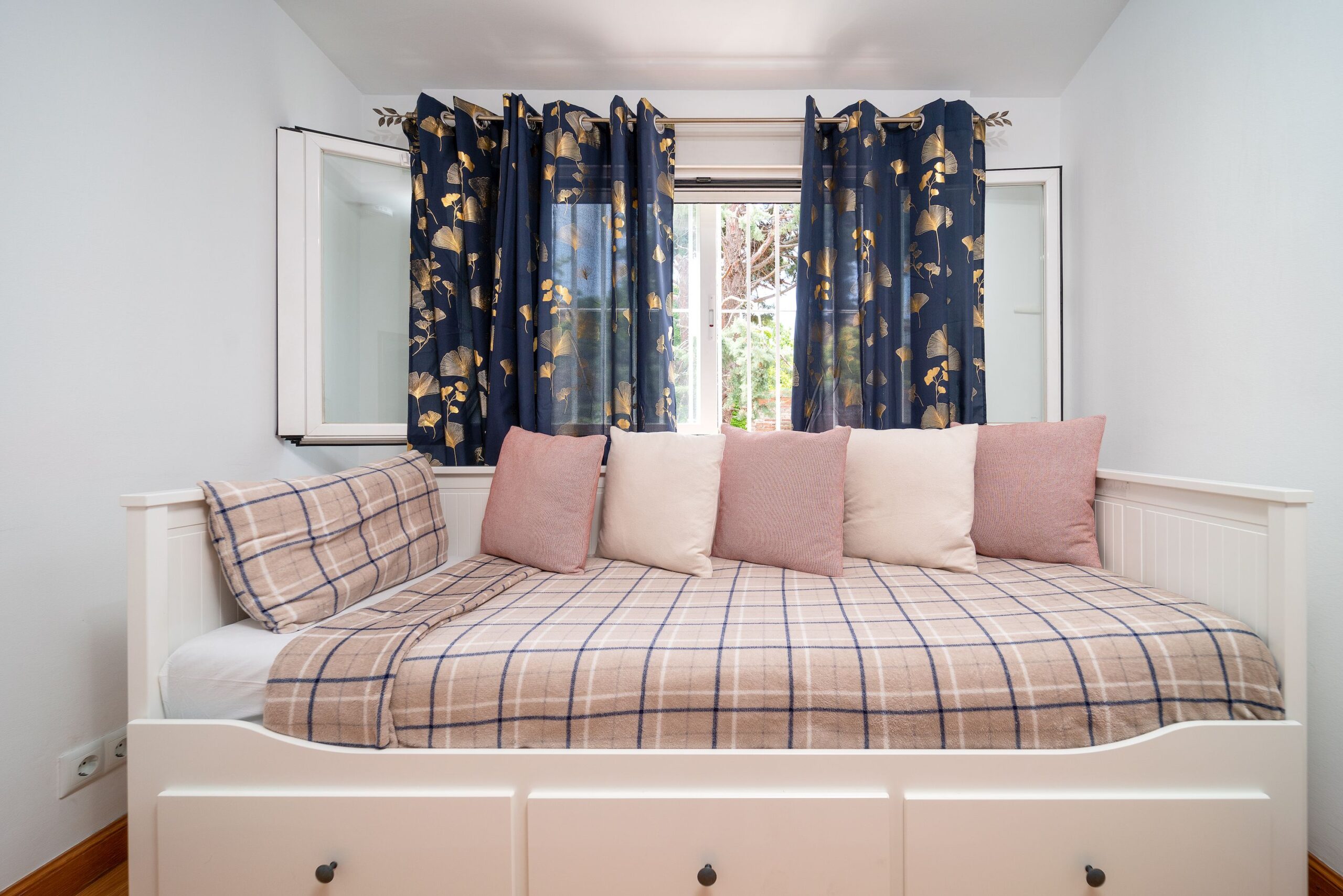 Cama tipo trineo blanca con almacenamiento; ropa de cama en cuadros beige y azul, cojines rosas y beige; cortinas azul marino con patrón; habitación juvenil luminosa y ordenada.