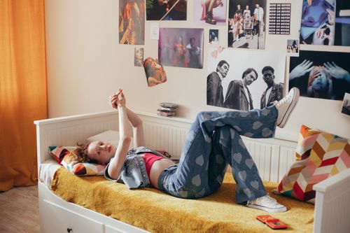 Chica adolescente recostada en la cama, piernas en el aire, mirando su móvil; habitación con paredes decoradas y colores vivos, cortina naranja y cojines geométricos. colores para habitación adolescente.
