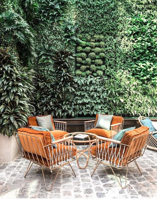 Conjunto de sillas de ratán natural con cojines naranjas y una mesa en un patio rodeado de plantas verdes.